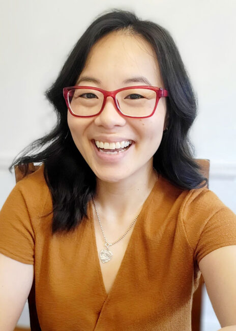 a photo of a smiling Jing-Jing Hu, she wears glasses and an orange shirt, Jing-Jing oversees the volunteer programs of Apex